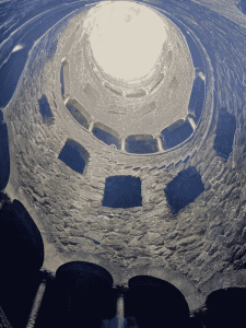 A view looking up from the bottom of the Initiation Well at the Quinta de Regaleira in Sintra Portugal. An ornate stone spiralling staircase leading up to a washed out sky.