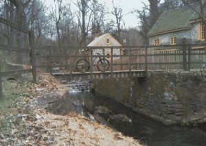 A dithered small bridge over a small walled-in creek with a yellow-detailed stone building behind. A mountain bike is posed on the bridge